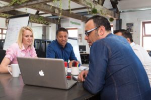 Three colleagues sit at a table reviewing content on a laptop
